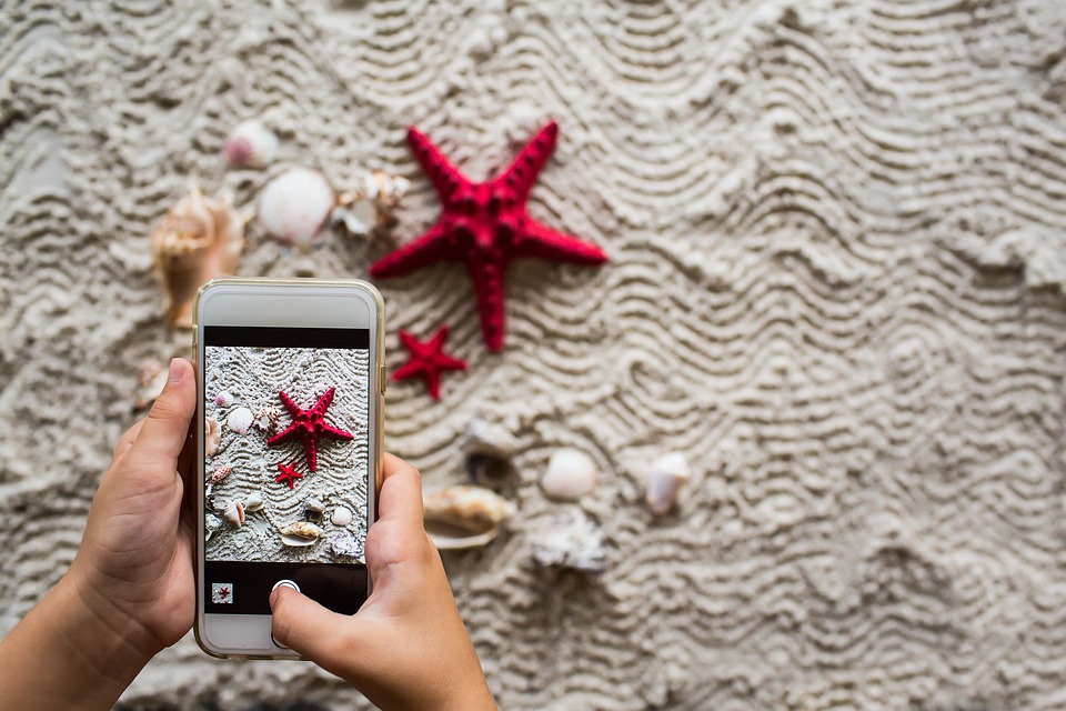 A mobile phone taking a picture of a starfish and shells on a white beach